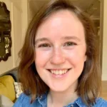 Portrait of Caroline Swift Holden smiling, wearing red glasses with short dark curly hair and a black sleeveless top.
