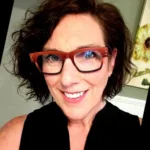Portrait of Tanya Coller smiling, with shoulder-length light brown hair, wearing a denim shirt in a warmly lit indoor space.